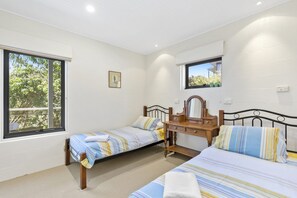 A bedroom with two single beds, a wooden dresser, a mirror, and a window with a view of trees. - Fernview (Lorne)