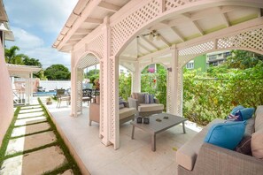 En overdækket udendørs terrasse med pool, siddeområde og udsigt til en grøn bygning.