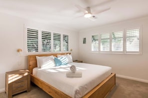 Une chambre à coucher avec un lit en bois, du linge de lit blanc, un ventilateur de plafond et des fenêtres équipées de stores.