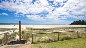 Ein Strand mit einer Holzpromenade, die zum Ufer führt, einer Rasenfläche im Vordergrund und einem klaren Himmel mit Wolken.