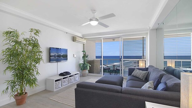 Ein modernes Wohnzimmer mit grauem Sofa, einem Flachbildfernseher, einem Deckenventilator und einem großen Fenster mit Blick auf den Ozean.
