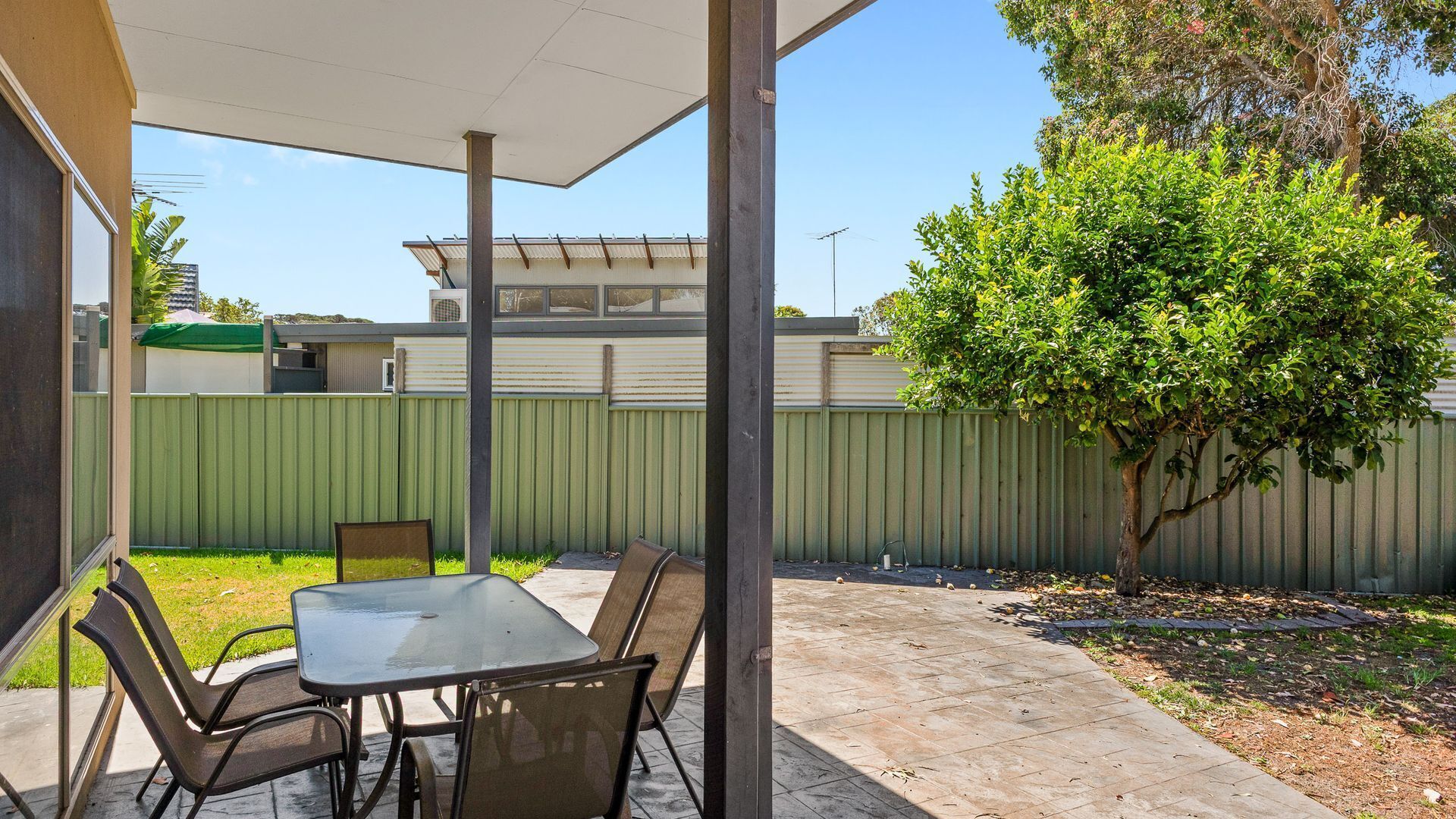 An outdoor patio with a table and chairs, a covered area, a tree, and a green fence.