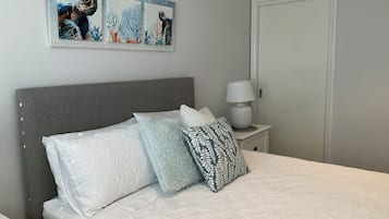 A neatly made bed with patterned pillows, a grey headboard, a wall-mounted artwork of sea turtles, and a bedside table with a lamp.