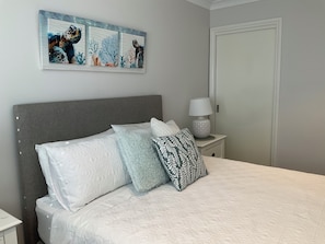 A neatly made bed with patterned pillows, a grey headboard, a wall-mounted artwork of sea turtles, and a bedside table with a lamp.
