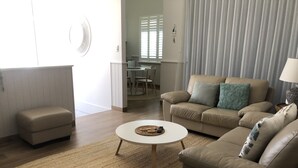 Un salon avec un canapé beige, une table basse ronde et un petit pouf. Au fond, on aperçoit un coin repas avec une table et des chaises.