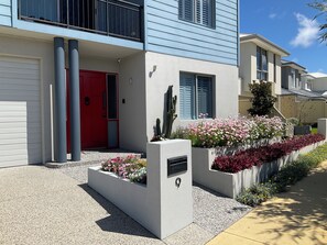 Een modern herenhuis met twee verdiepingen, een rode deur, een balkon en een verzorgde tuin met diverse planten en bloemen.