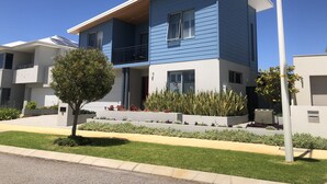 Un immeuble résidentiel moderne à deux étages, doté d’un toit plat, d’un balcon à l’étage supérieur et d’un jardin bien entretenu à l’avant, avec un petit arbre et des arbustes bas.