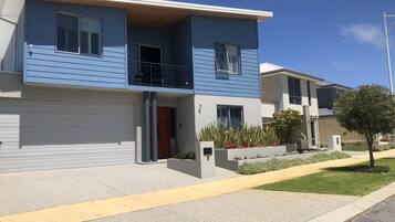 A two-story modern residential building with a red door, a balcony, and a small front yard with plants.