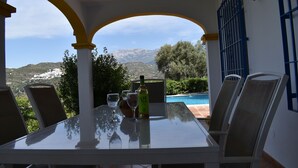 Ein Tisch mit Glasplatte, zwei Stühle, eine Flasche und zwei Gläser in einer überdachten Außenbereich mit Blick auf einen Berg und ein Schwimmbad.
