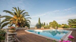 A pool with a wooden deck, surrounded by potted plants and flowers, with a view of trees and a clear sky.