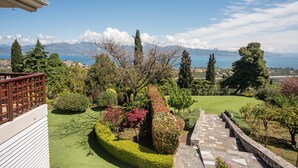 A landscaped garden with a stone pathway, a variety of shrubs and trees, and a view of the sea and mountains in the background.