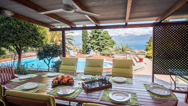 A covered outdoor dining area with a pool view, wooden tables, yellow chairs, and a table set with plates, glasses, and fruit.