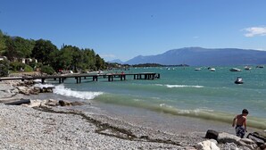 Una playa rocosa con un muelle que se adentra en el agua, barcos y montañas al fondo.