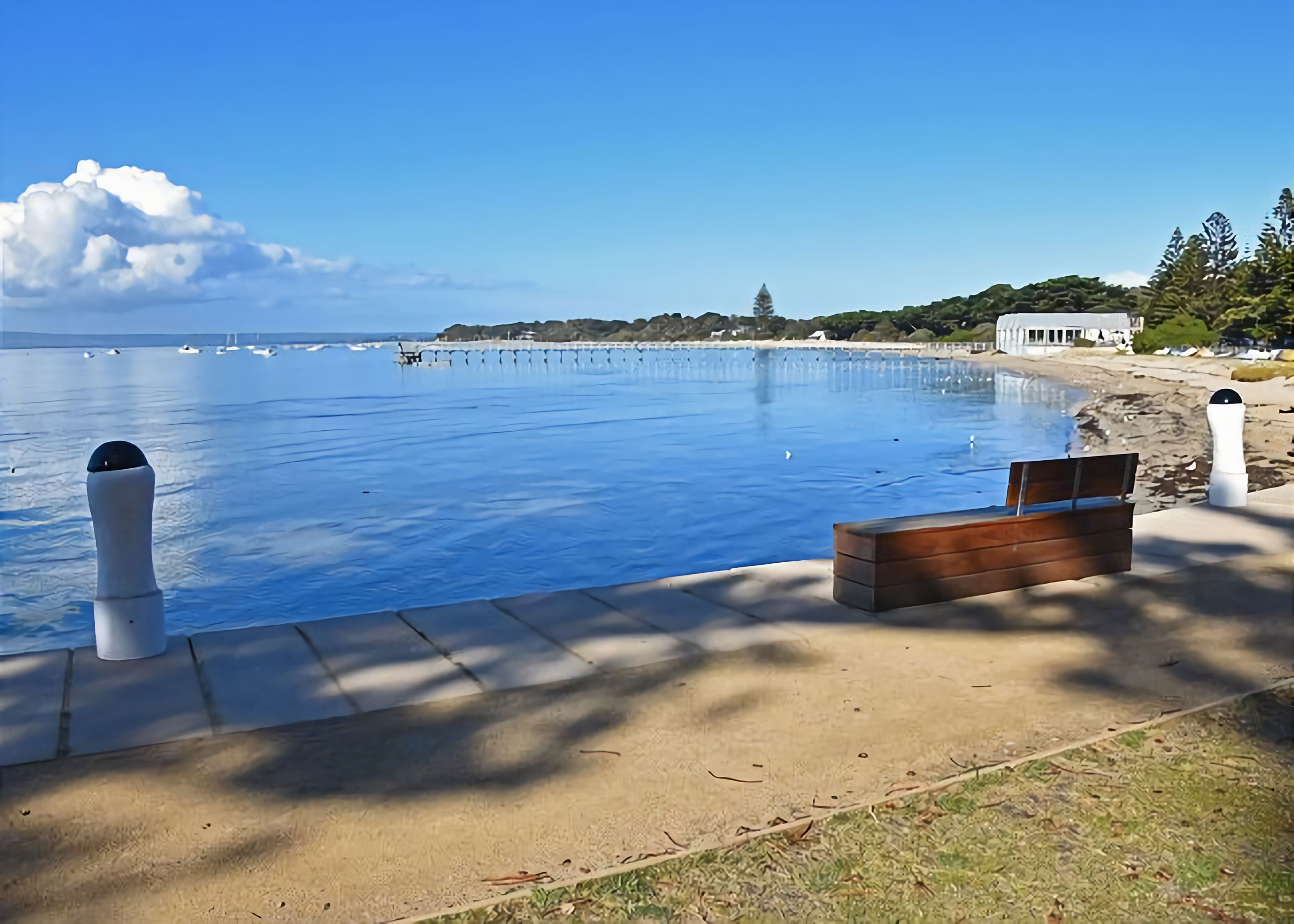 A clear view of a swimming pool with a wooden bench and bollards, a sandy area, and a distant shoreline with buildings.