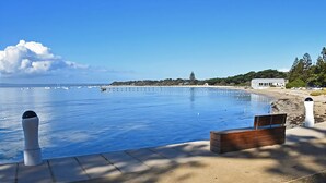 On aperçoit clairement une piscine, avec un banc en bois et des bollards, entourée d’une zone sablonneuse et d’un bâtiment en arrière-plan.
