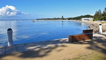 A clear view of a swimming pool with a wooden bench and bollards, a sandy area, and a distant shoreline with buildings.