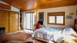 A bedroom with a wooden ceiling, a bed with a floral bedspread, a desk with a chair, a bookshelf, and a sliding glass door leading to a balcony.