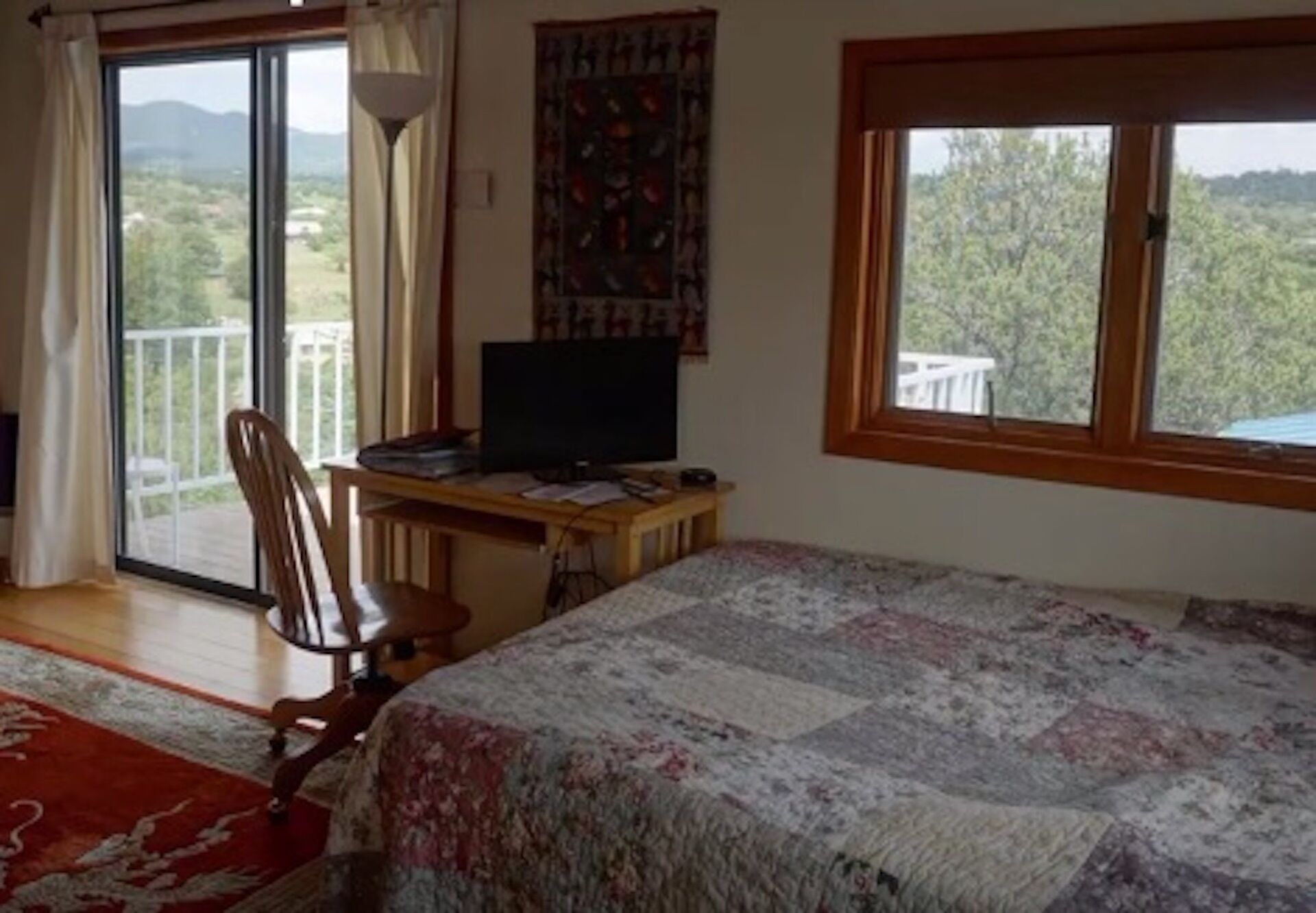 A bedroom with a bed, a desk with a computer, a chair, a window with a view of greenery, and a balcony.