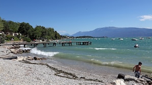 A rocky beach with a pier extending into the water, boats, and mountains in the background. - Three-room villa direct to the beach, 6 beds garden pool  (Manerba del Garda)