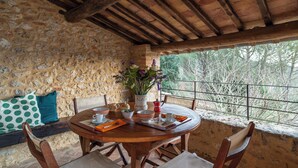 Un mur de pierres apparentes, des poutres en bois au plafond, une table ronde en bois entourée de chaises, un vase de fleurs et une vue sur les arbres à travers la rambarde d’un balcon.