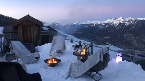 A cozy outdoor seating area with a fire pit, covered chairs, and a wooden structure, overlooking a snowy mountain range.
