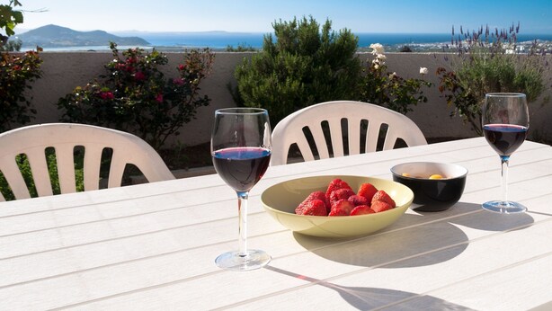 Una mesa con dos copas de vino y un bol de fresas, frente a una vista panorámica del mar y las montañas.