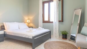 Una cama bien hecha con ropa de cama y toallas blancas, una lámpara de noche, una planta en maceta, un espejo y una alfombra redonda de tejido en una habitación con paredes verde claro.