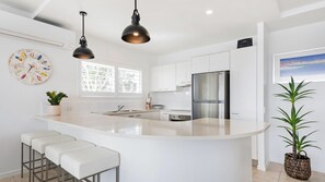 A modern kitchen with white cabinetry, a stainless steel refrigerator, a large island with bar stools, pendant lighting, and a potted plant. - LOCATION DOESN'T GET ANY BETTER THAN THIS (Noosaville)