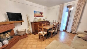 Een woonkamer met een open haard, een eettafel met stoelen, een televisie op een plank en een schilderij aan de muur.