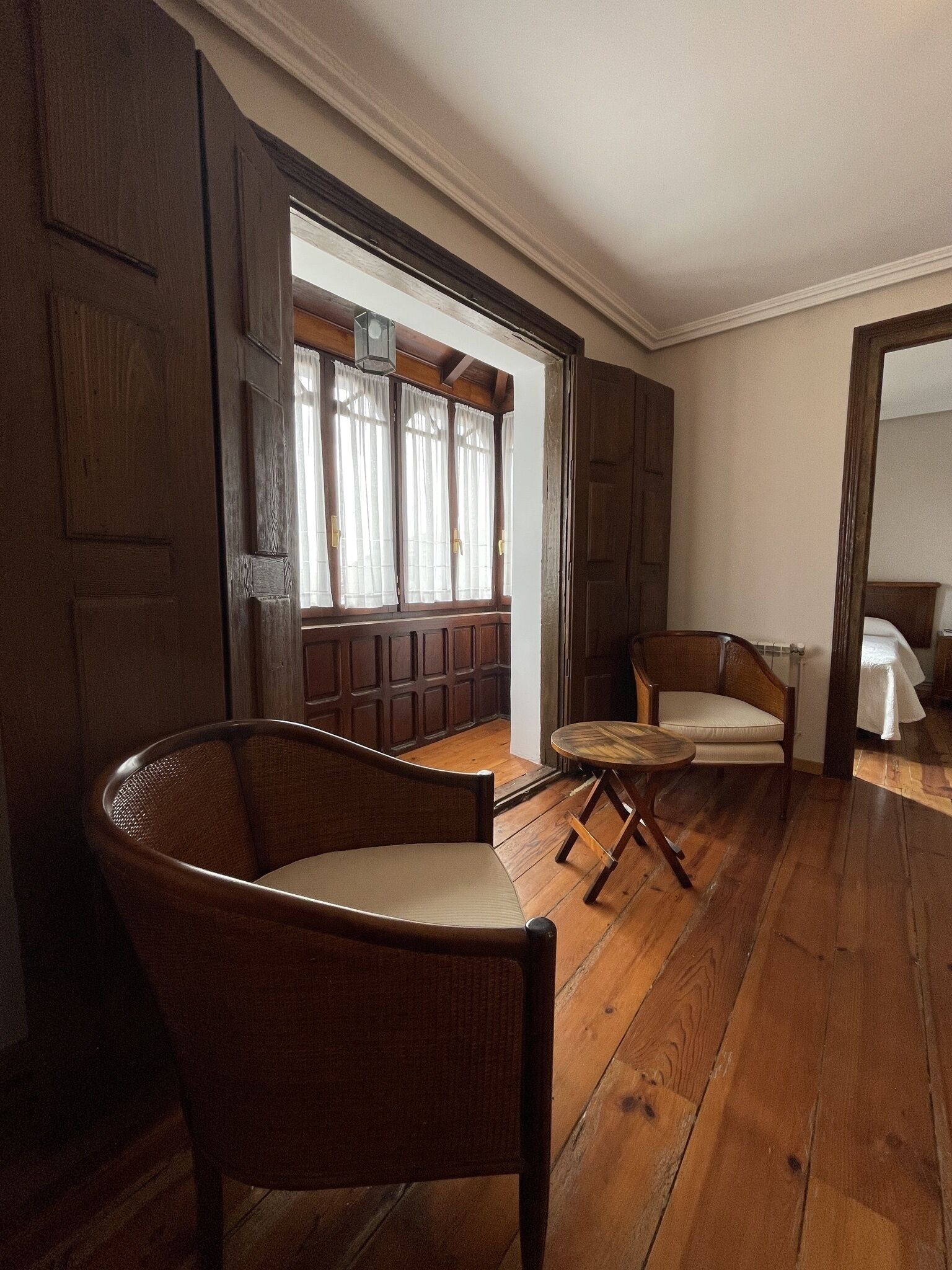 Una habitación con suelo de madera, una silla de mimbre, una pequeña mesa redonda y un banco con cortinas.