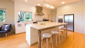Uma cozinha moderna com ilha central, banquetas, geladeira de aço inoxidável e vista para o exterior.