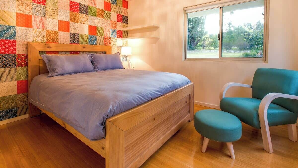 A bedroom with a wooden bed, a quilted wall, a teal chair, and a window with a view of greenery.
