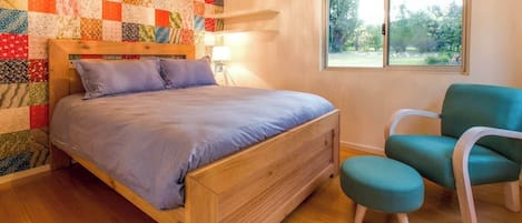 Un dormitorio con cama de madera, pared acolchada, una silla color turquesa y ventana con vista a follaje.