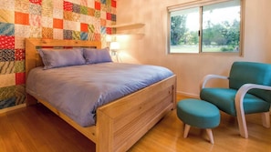 A bedroom with a wooden bed, a quilted wall, a teal chair, and a window with a view of greenery.