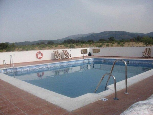 Ein rechteckiger Swimmingpool mit einer gefliesten Kante, umgeben von einer weißen Mauer. Auf beiden Seiten befinden sich Metallleiter zum Pool, und an der Mauer ist ein roter Rettungsring befestigt. Der Pool wird von Hügeln und einem klaren Himmel im Hintergrund begleitet.