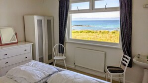Ein Schlafzimmer mit Bett, Schminktisch, Stuhl, Heizkörper, Fenster mit Meerblick und einem Spiegel auf dem Schminktisch.