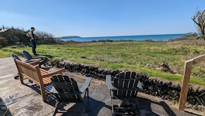Eine Terrasse mit Holzstühlen und einem Tisch mit Blick auf ein saftiges Grasfeld und das Meer.