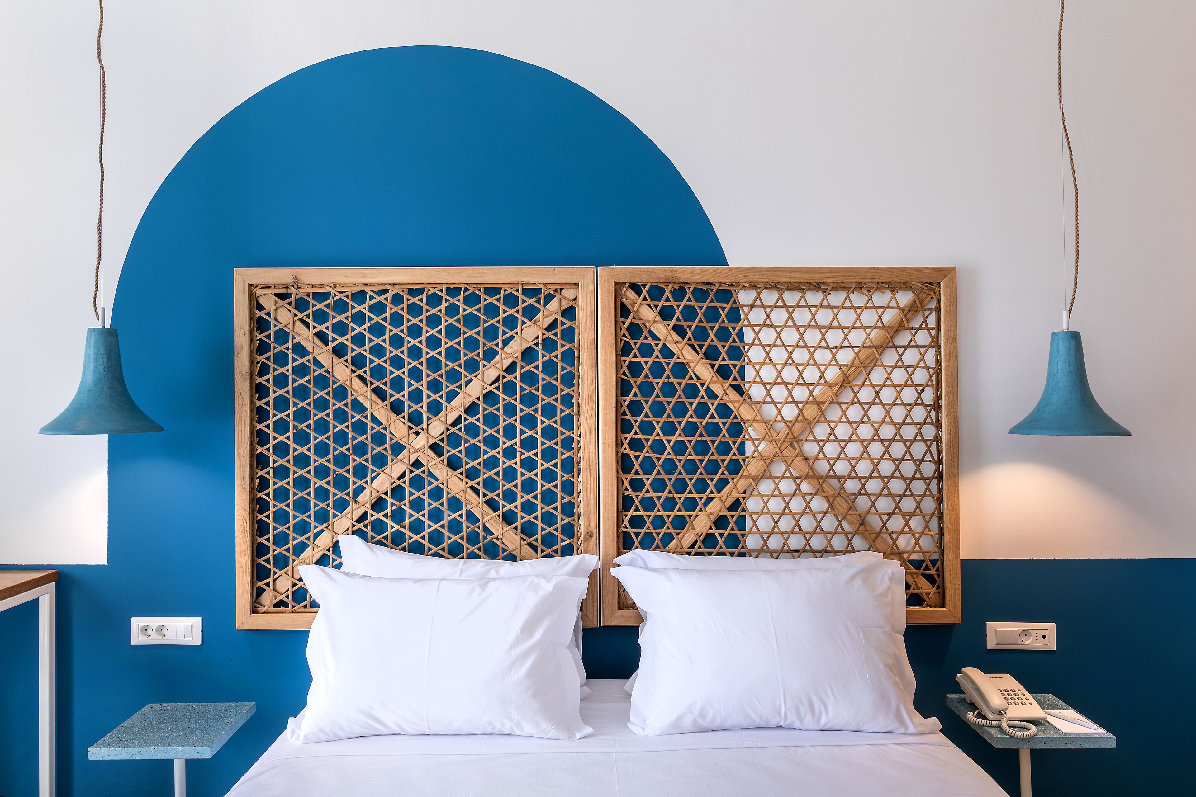 A hotel room with a bed, two white pillows, a wooden lattice headboard, a blue wall, and a small table with a phone.