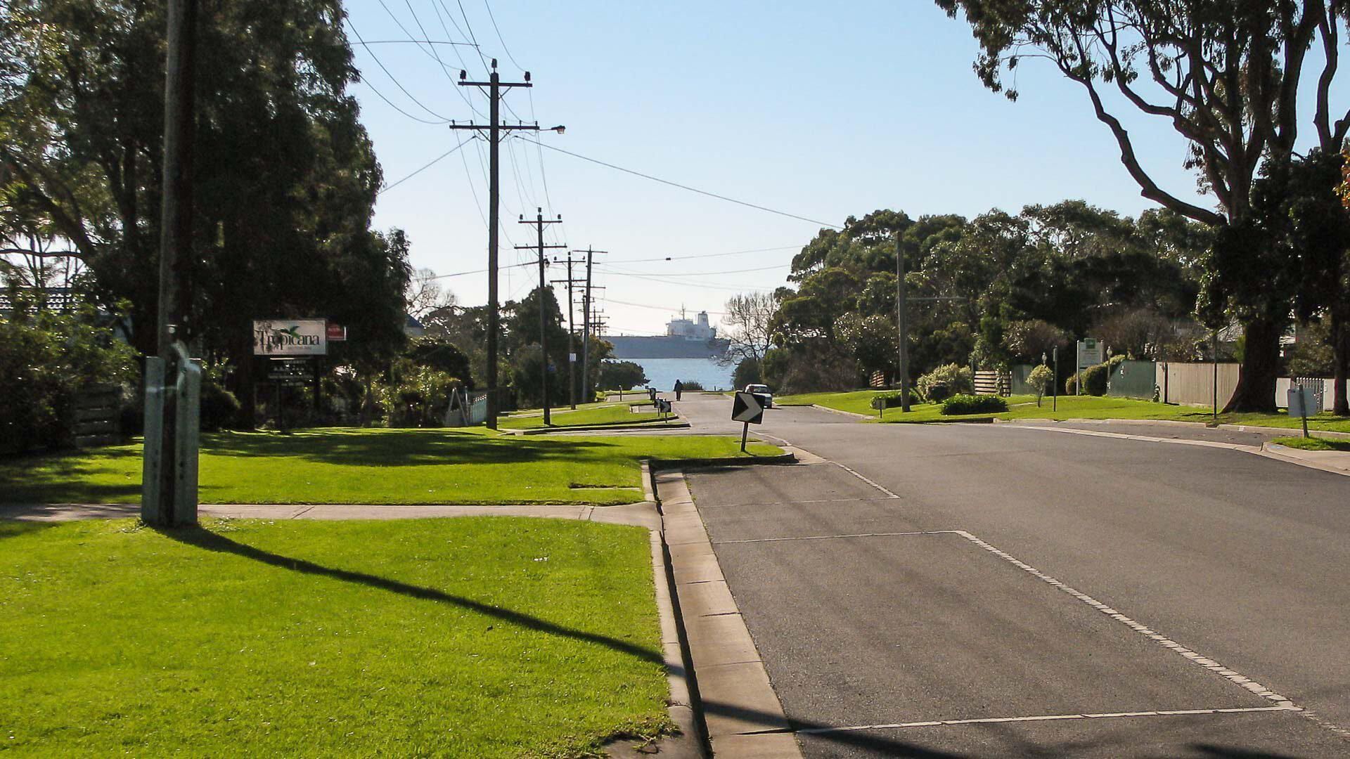 A quiet suburban street with a grassy median, utility poles, and a distant view of a body of water.