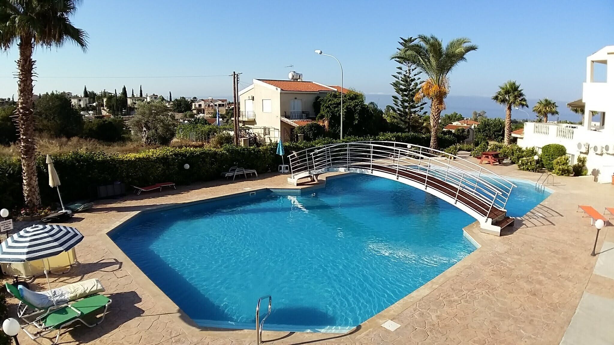 A swimming pool with a curved bridge, surrounded by palm trees and a clear view of the sea.