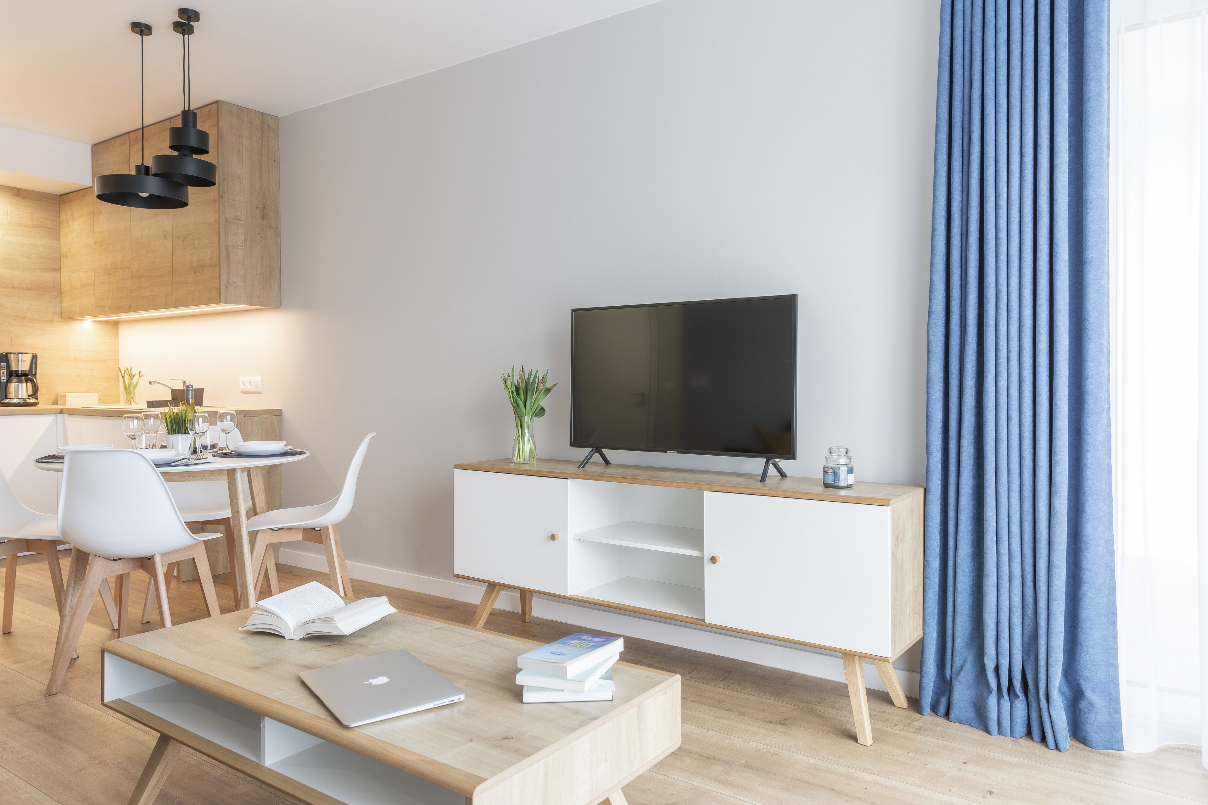 Ein modernes Wohnzimmer mit Flachbildfernseher, Holzmöbeln und einem Essbereich mit weißen Stühlen und einem Tisch.