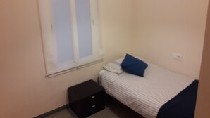 Ein kleines Zimmer mit einem Einzelbett, einem Fenster, einem Nachttisch und einer Steckdose.