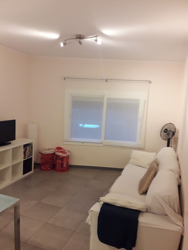 A living room with a white sofa, a TV on a stand, a window with blinds, and a ceiling fan.