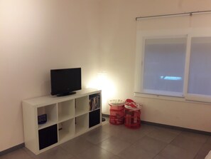 Una habitación con televisión sobre una estantería blanca, una pantalla de proyector y una ventana con vistas.