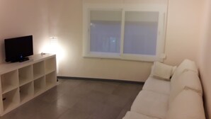 Ein Zimmer mit einer weißen Couch, einem Fernseher auf einem Schrank und einem Fenster mit Jalousien.