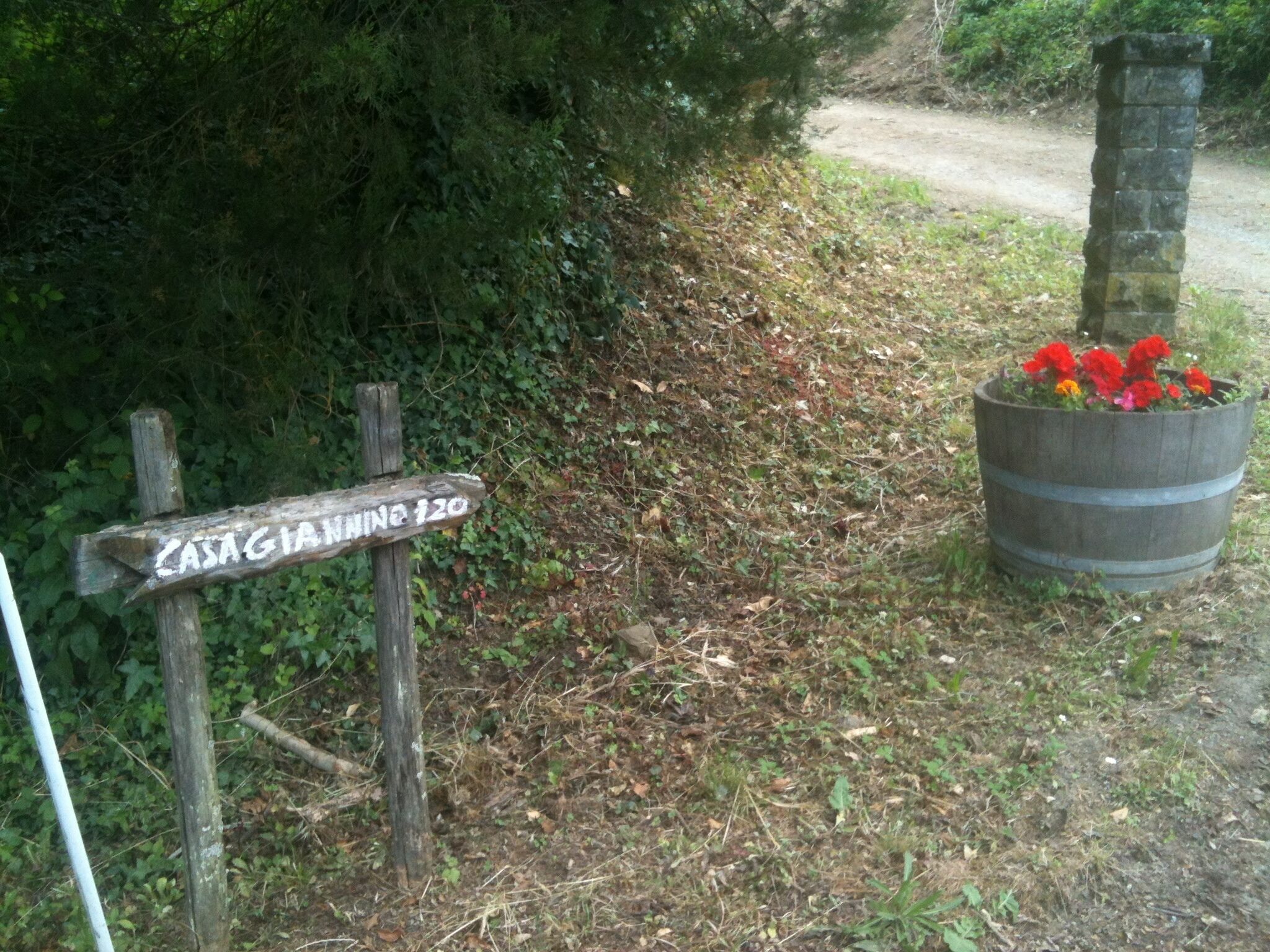 A wooden signpost with 'CASAGIANNA 120' written on it, a stone pillar, and a barrel with flowers.