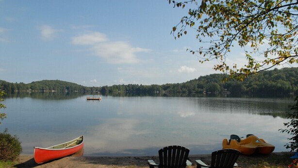 A calm lake with a canoe and two chairs on the shore, surrounded by trees and hills. - Muskoka Cottage with a Sandy Beach (Baysville)