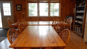 Une table à manger en bois, des chaises, une bibliothèque et une fenêtre donnant sur les arbres.