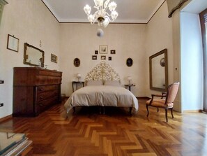 Une chambre avec un parquet en bois disposé en motif de chevron, un grand lit agrémenté d’une tête de lit décorative, une commode, une chaise et un miroir au mur.