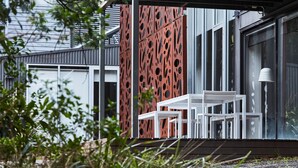 A modern outdoor seating area with a white table and chairs, a red perforated wall, and a glass door leading to a building.